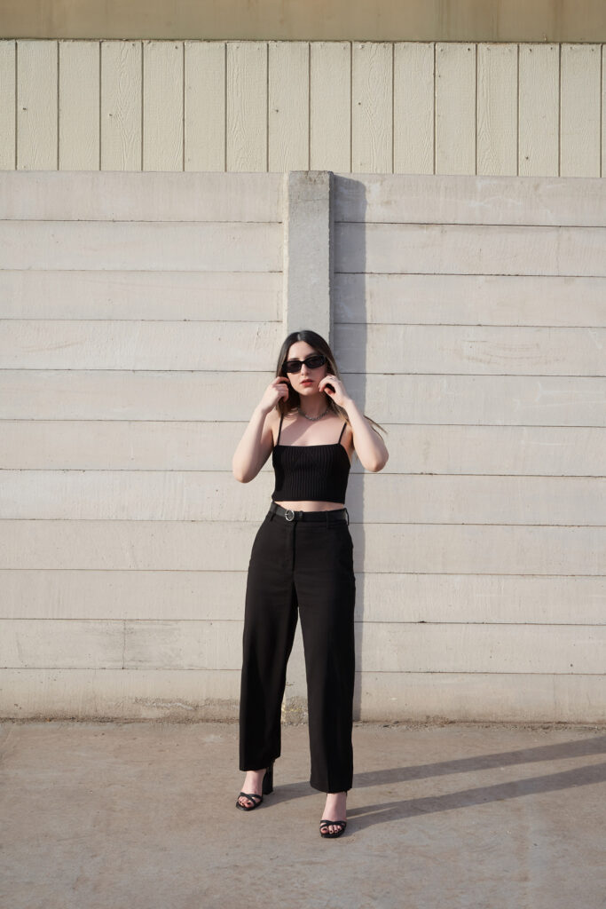 Gabriela Inzunza for Avice a modern minimalist look with a black tube top high-waisted black trousers and black strappy heels posed against a beige wooden wall.